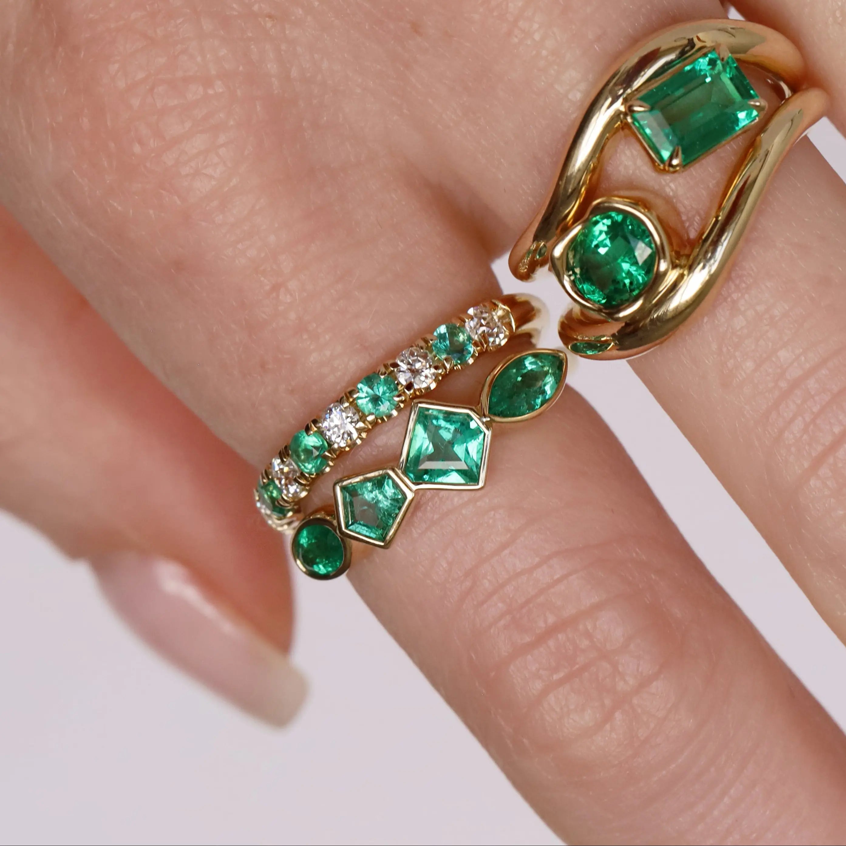 Gold rings with emerald green gemstones on a finger against a neutral background
