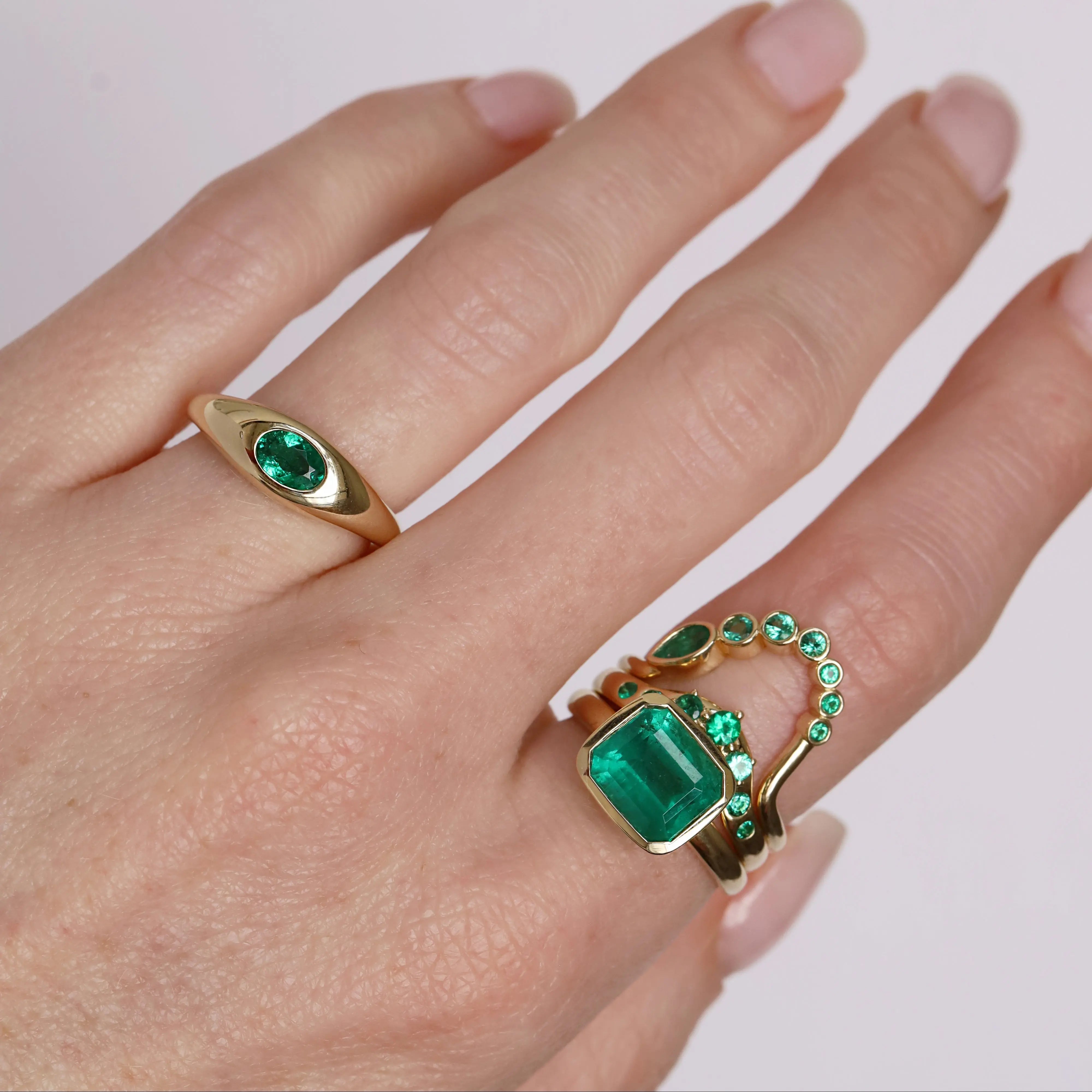 Hand wearing two gold rings with green gemstones on a light background