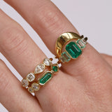 Gold ring with emerald and diamond stones on a finger against a white background