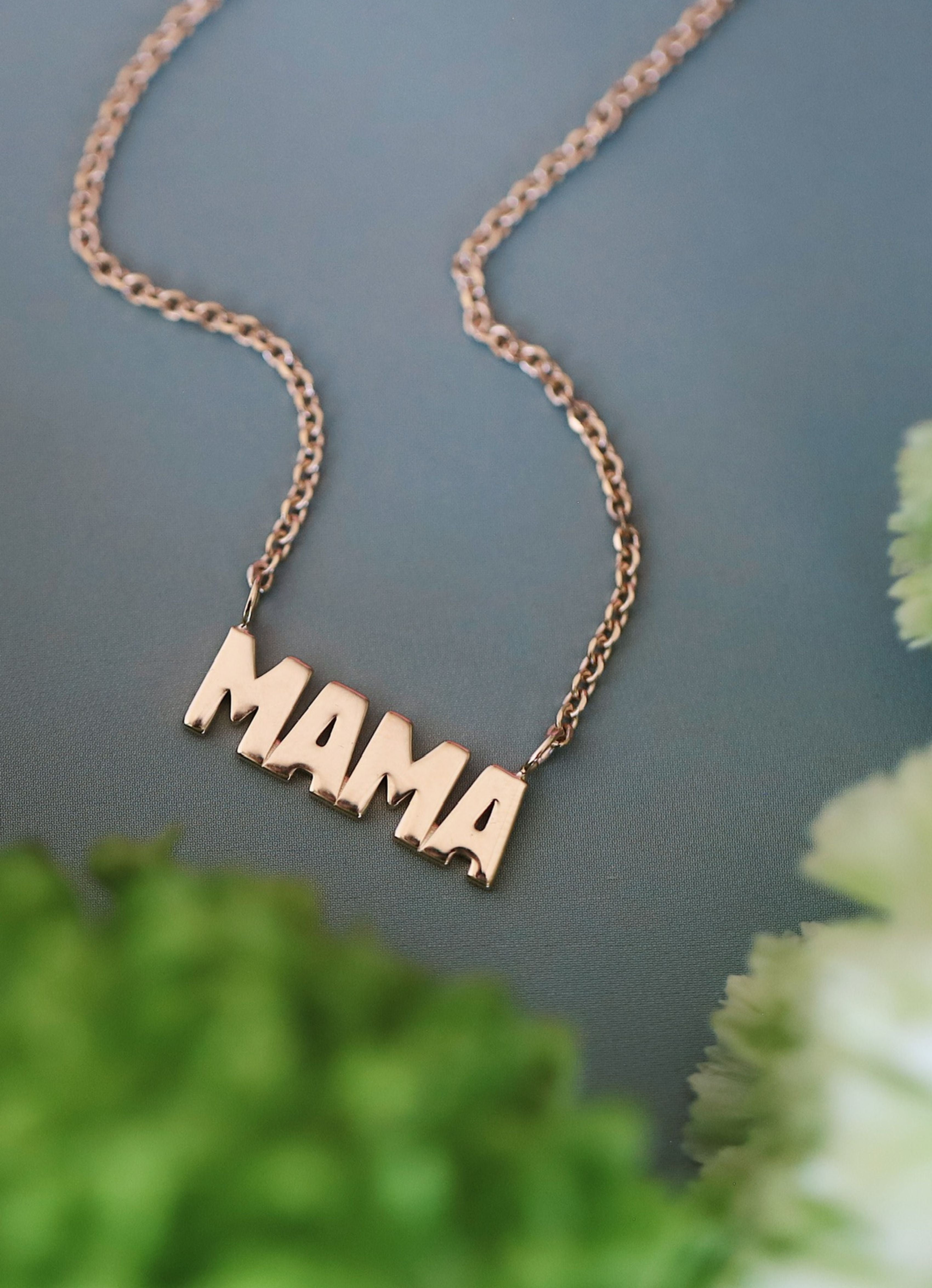 Gold mama necklace against a blue background with greenery/flowers peeking through in the corner