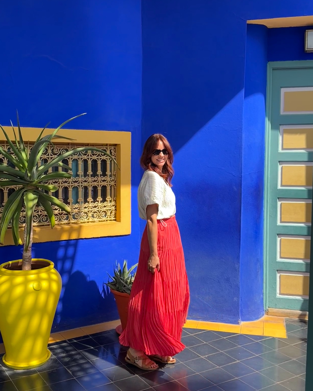 Woman in a white top and flowing red skirt standing against a cobalt blue wall with yellow planters in Marrakech