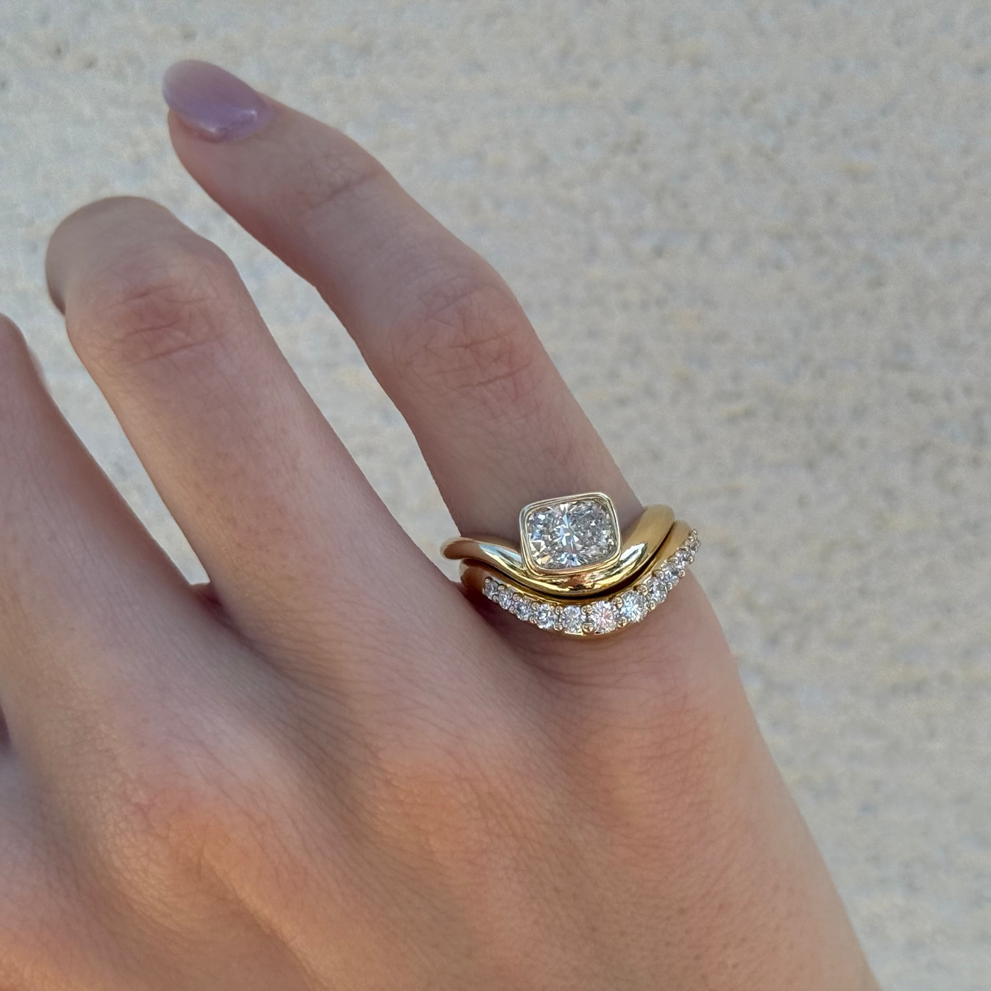 A close-up of a hand wearing a gold ring with a square-cut clear center stone and a curved pavé band, set against a light textured background.