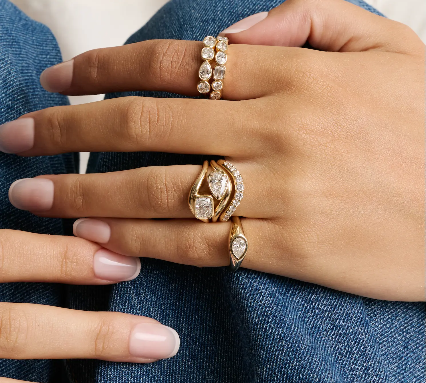 A close-up of a hand wearing multiple gold rings with clear gemstones, resting on blue denim fabric.