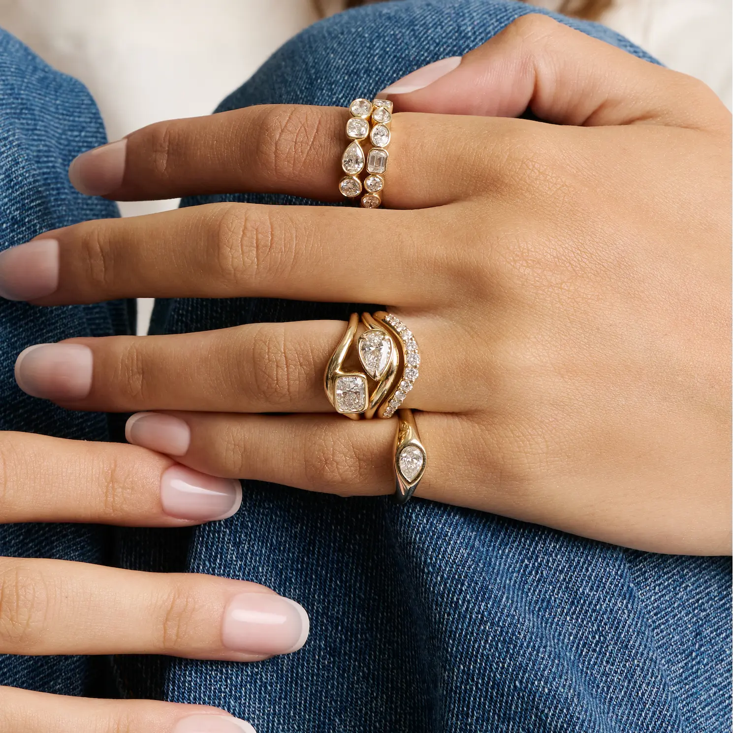 A close-up of a hand wearing multiple gold rings with clear gemstones, resting on blue denim fabric.