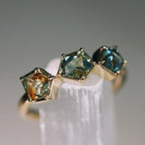 Gold ring with three green gemstones on a white stand against a gray background