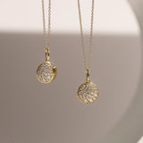 Two gold necklaces with round, diamond-studded pendants on a neutral background.
