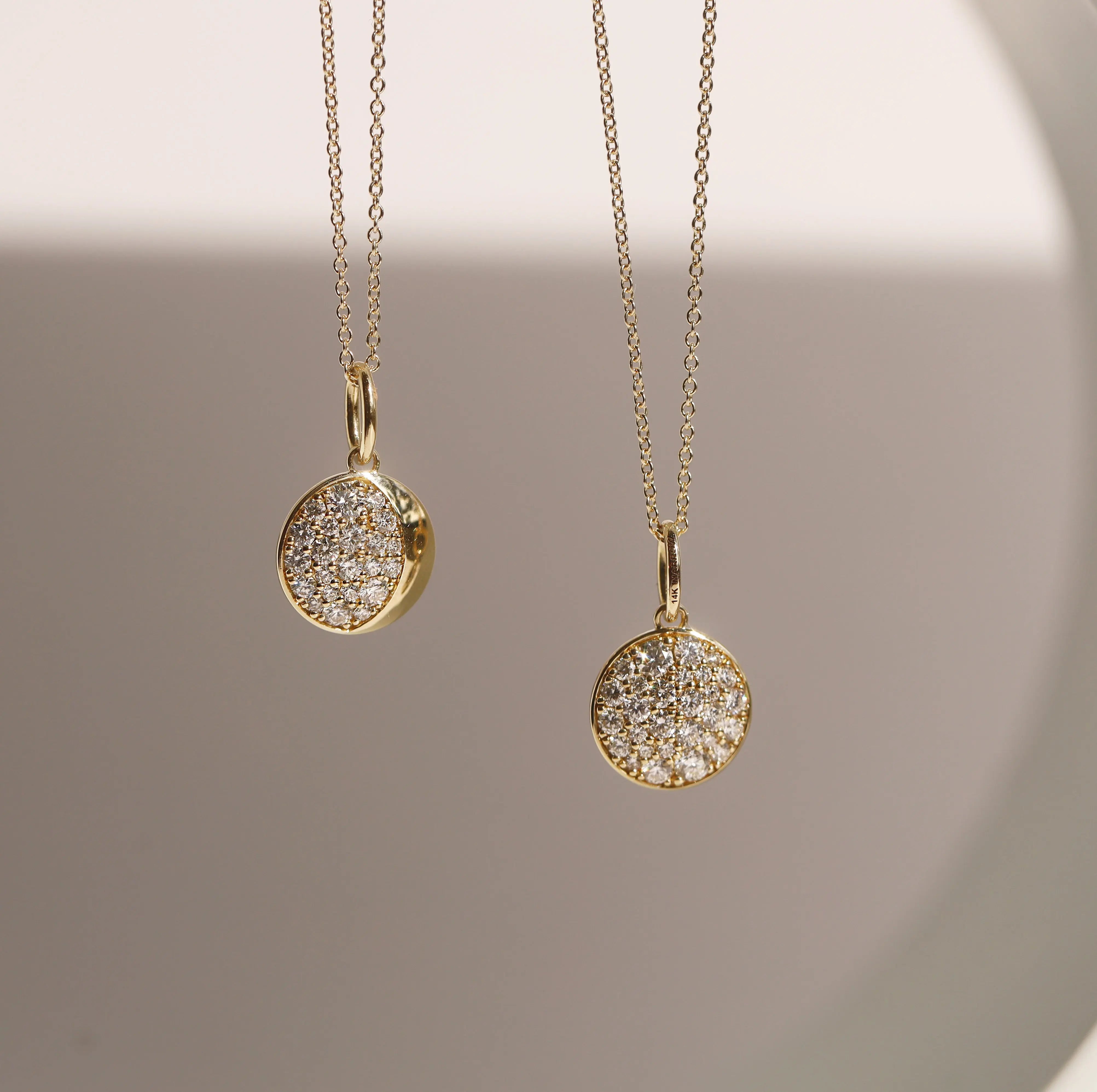 Two gold necklaces with round, diamond-studded pendants on a neutral background.