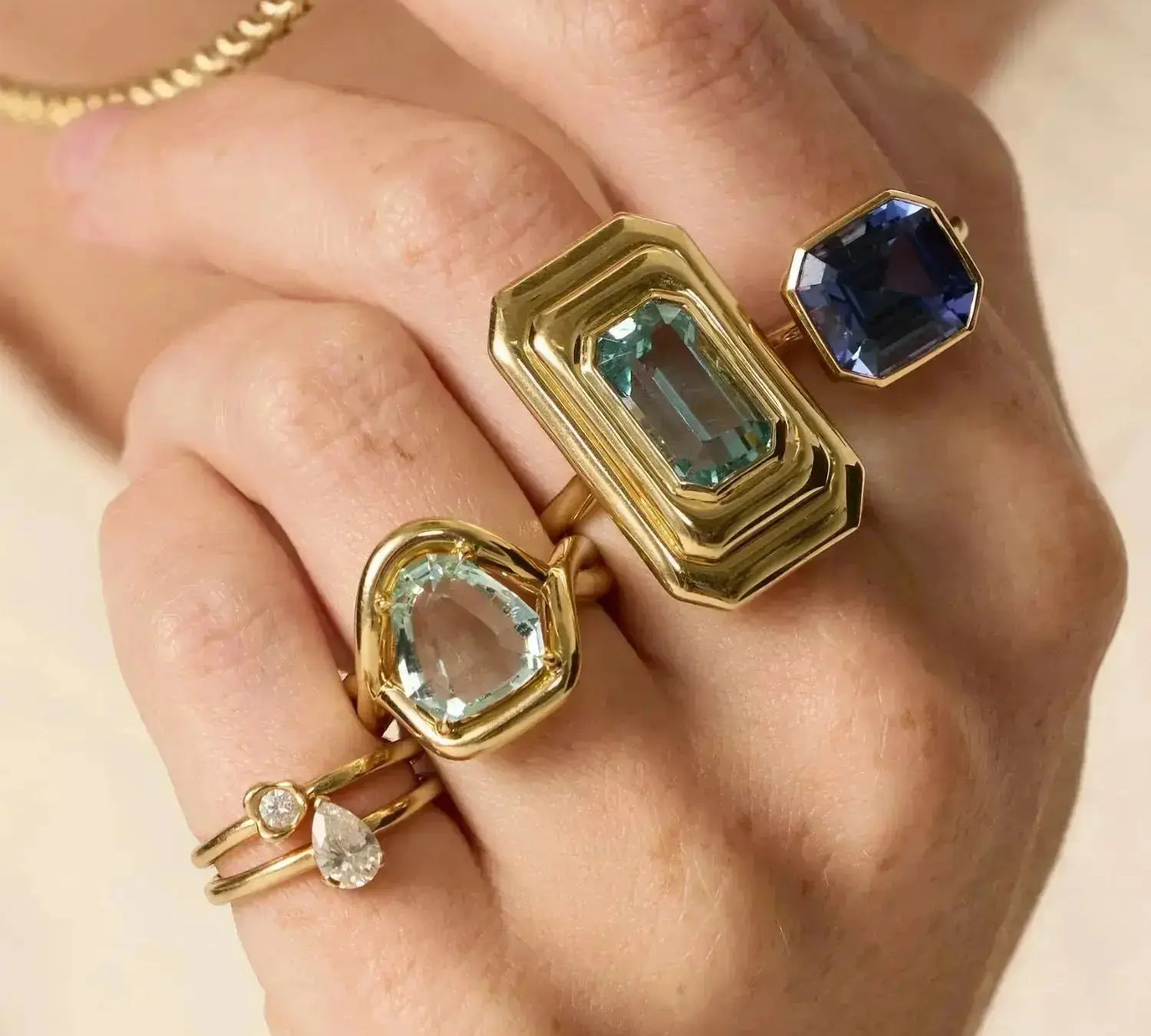Hand wearing three gold rings with gemstones on a neutral background