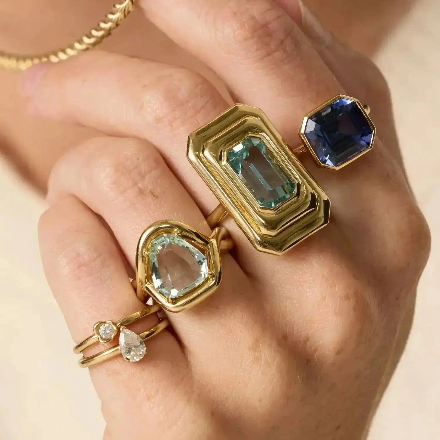 Hand wearing three gold rings with gemstones on a neutral background
