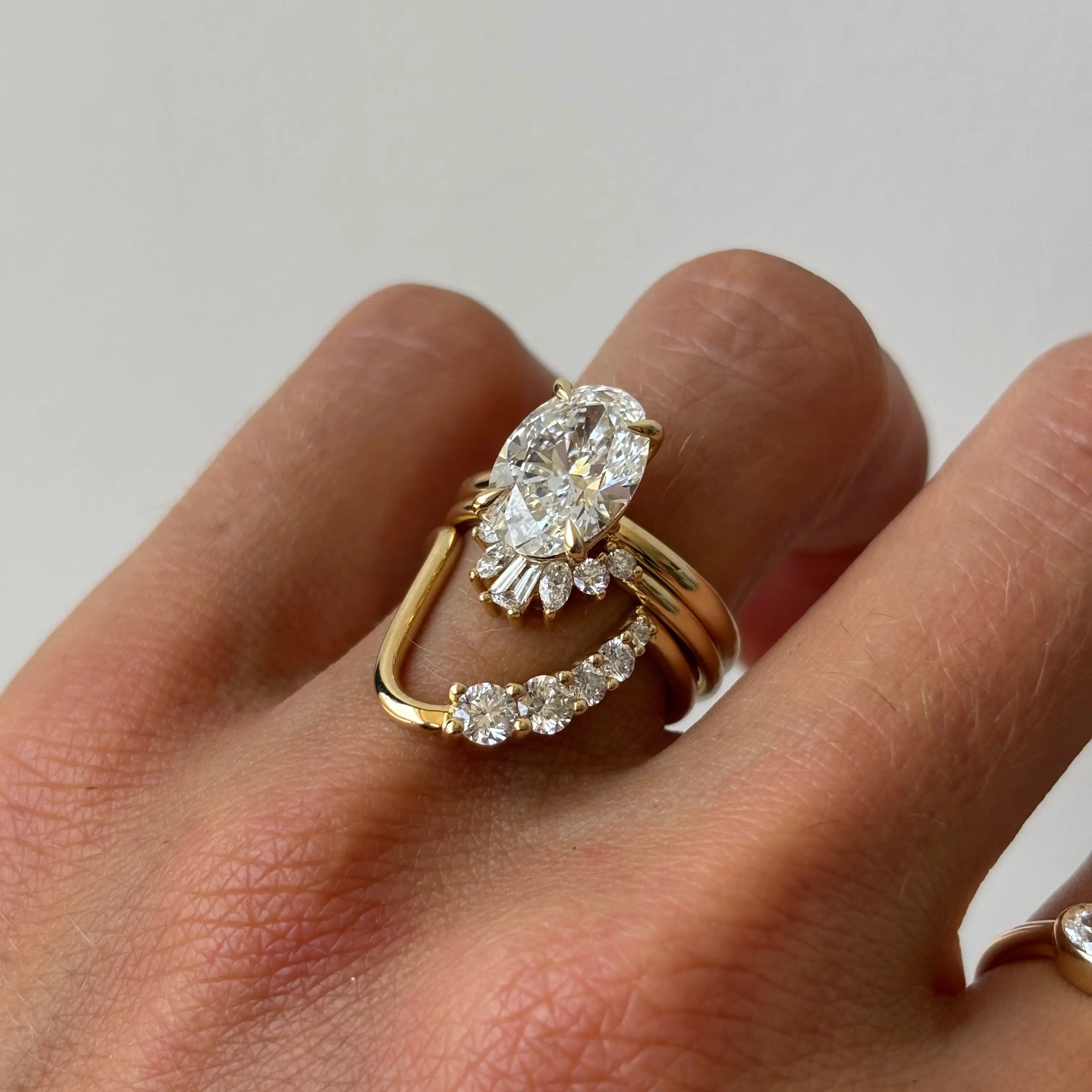 A close-up of a hand wearing a yellow gold ring set with a large oval diamond and smaller accent diamonds on a plain light background.