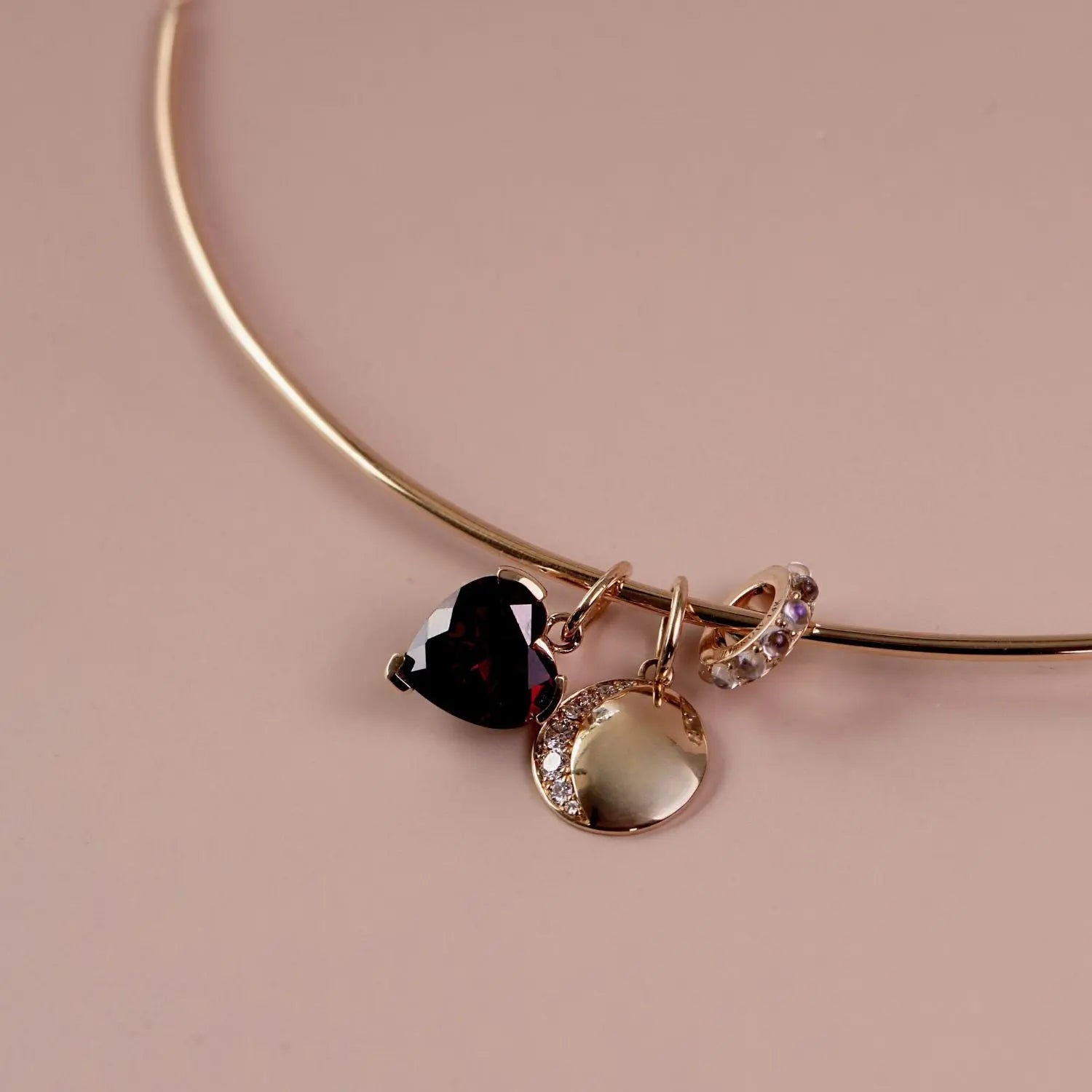 A gold collar necklace with three dangling charms: a dark red heart, a gold disc with moonstones and a small gold crescent with purple stones, set against a solid dusty pink background.