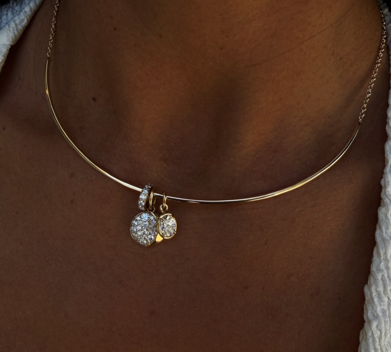 A close-up of a person's neck wearing a delicate gold bangle necklace with a small dangling charm covered in clear stones.