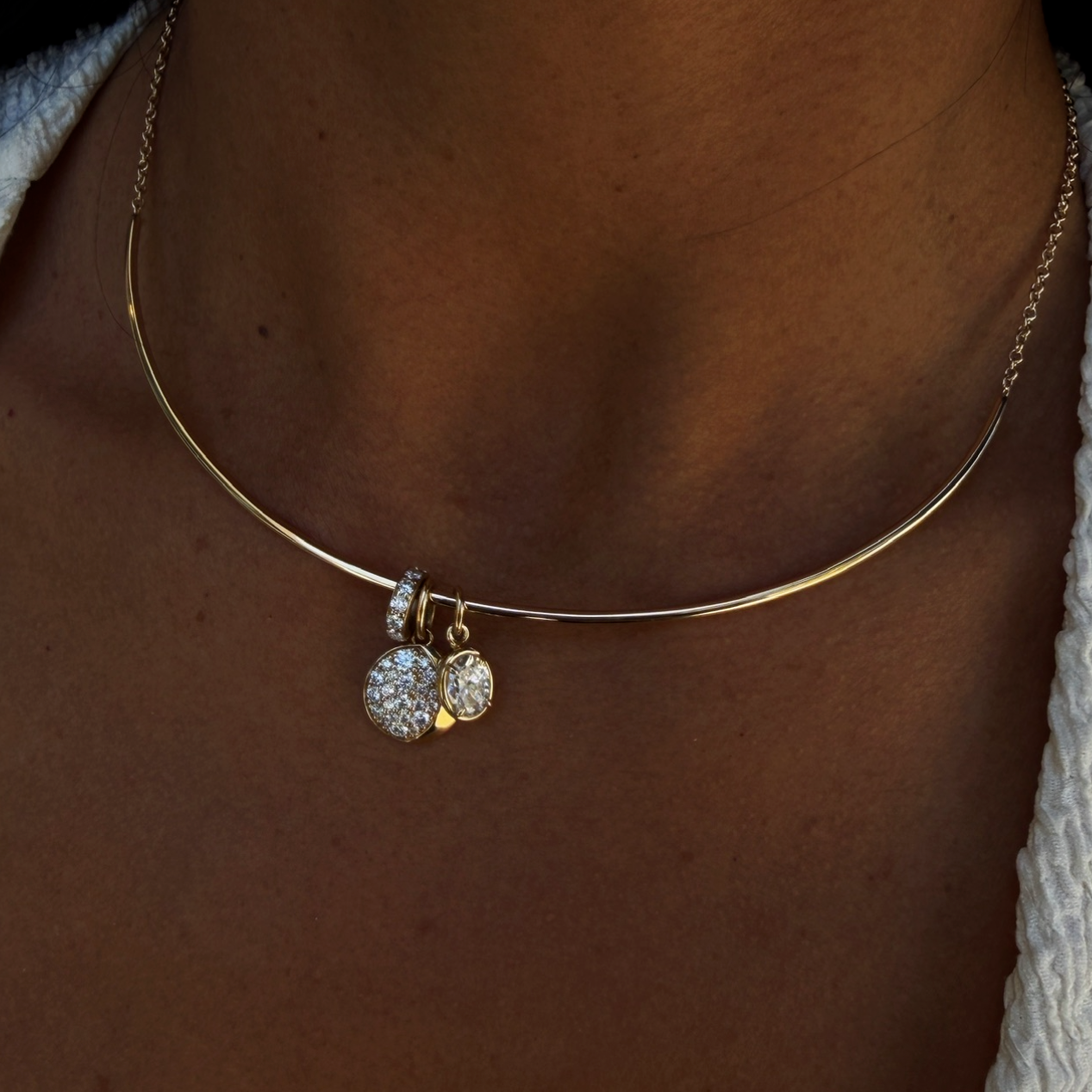 A close-up of a person's neck wearing a delicate gold bangle necklace with a small dangling charm covered in clear stones.