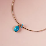 A gold collar necklace with a vibrant turquoise oval pendant and a small beaded accent, displayed on a soft pink background.