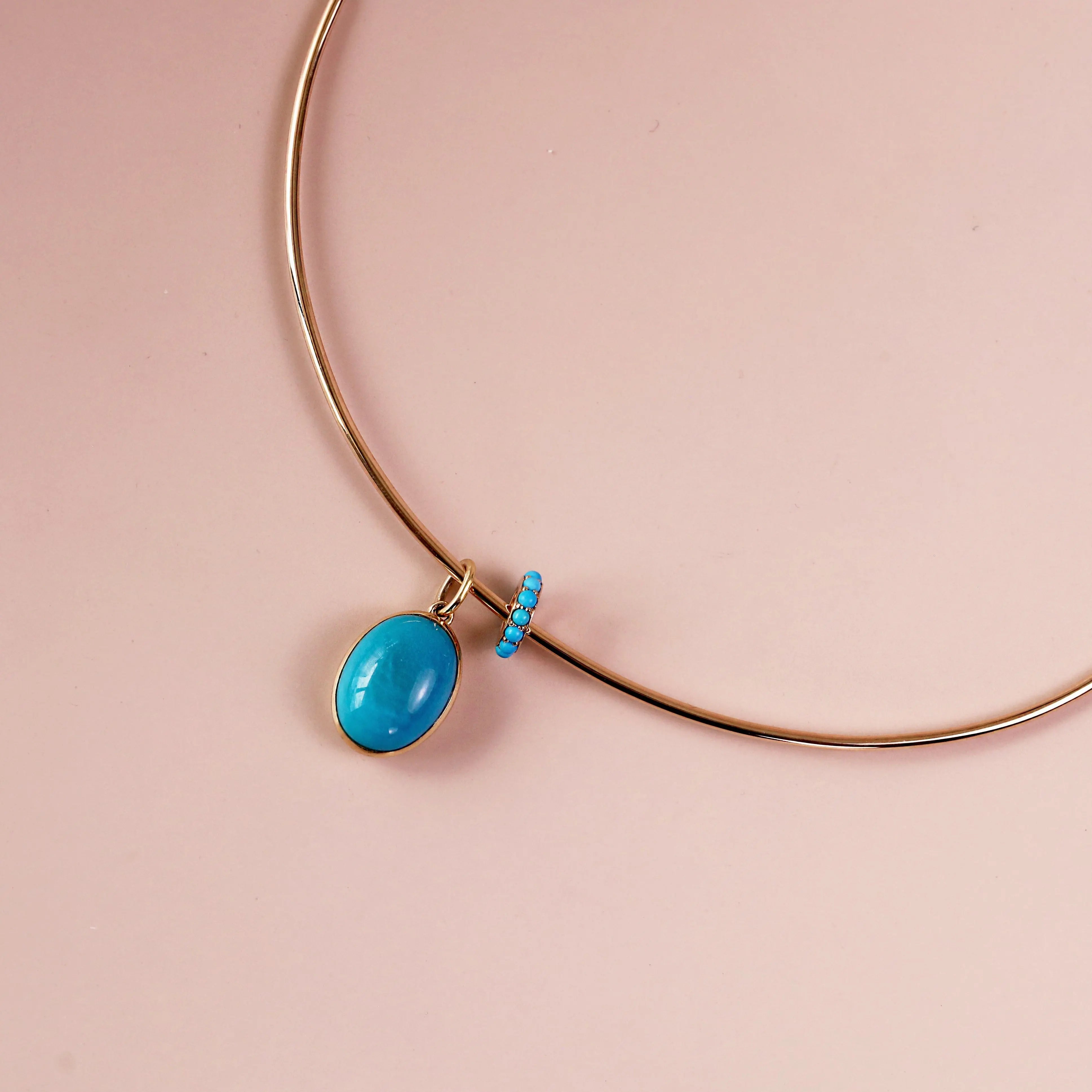 A gold collar necklace with a vibrant turquoise oval pendant and a small beaded accent, displayed on a soft pink background.