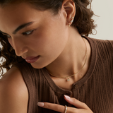 A close-up lifestyle shot of a woman wearing delicate gold jewelry, including a small gold necklace with a crystal pendant, a gold ring, and a gold ear cuff, paired with a brown ribbed knit top.