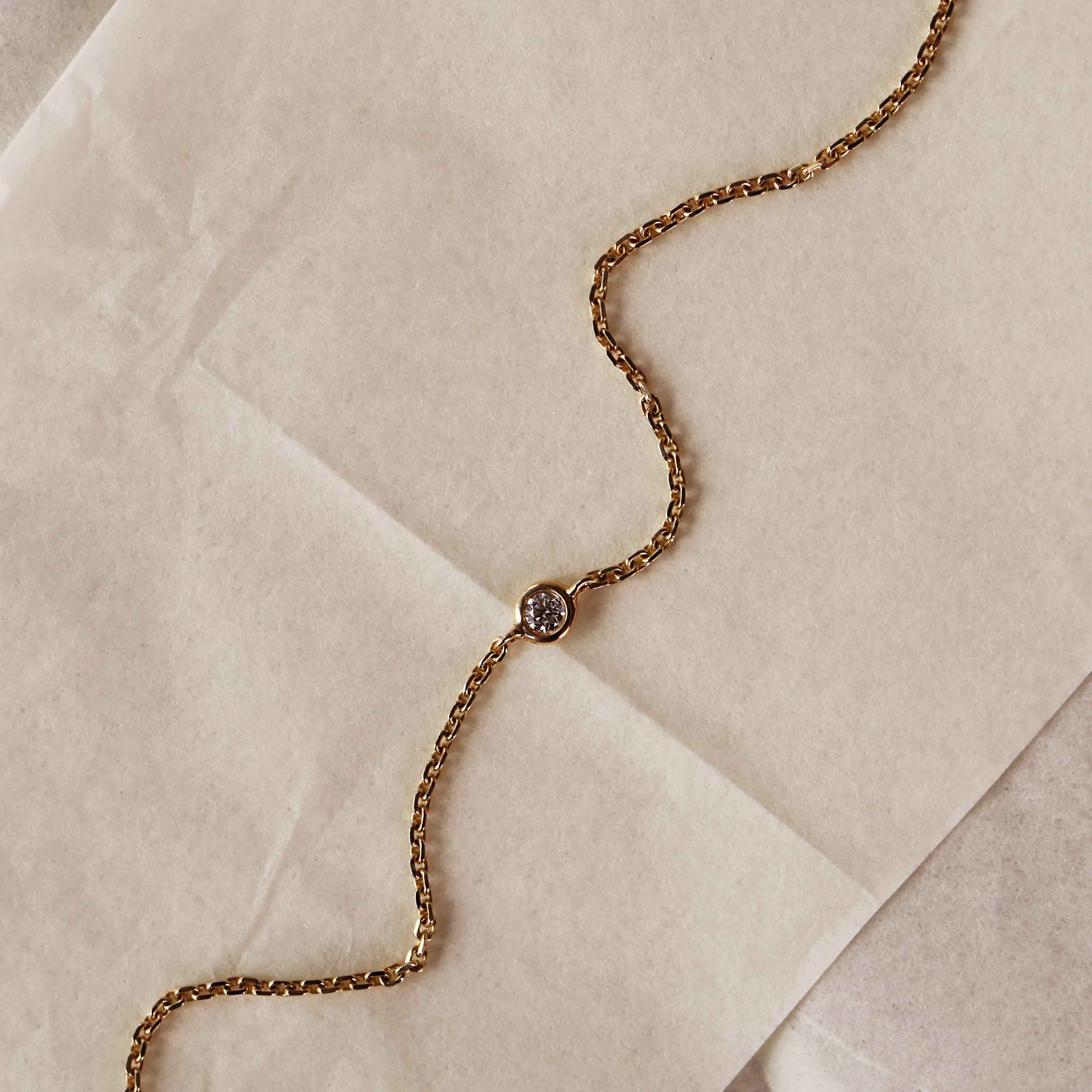 A close-up of a gold chain bracelet with a single round bezel-set diamond, laid diagonally across a textured beige surface.