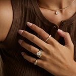A close-up lifestyle shot of a person wearing multiple gold rings with clear gemstones, including an engagement-style ring with a round center stone and a beaded accent band, along with a delicate gold necklace.