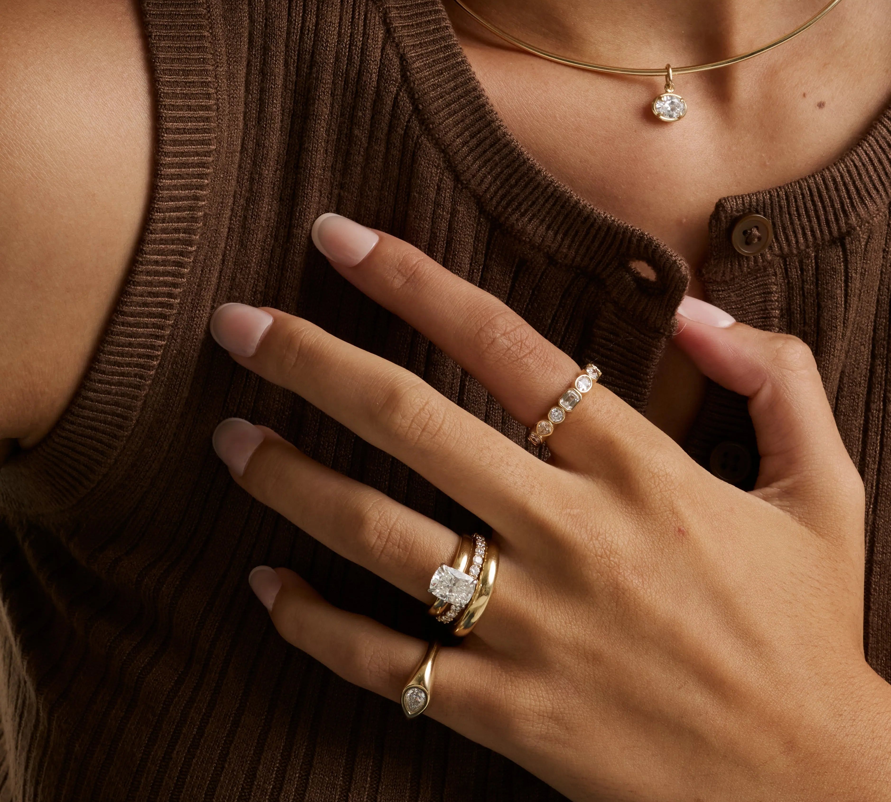 A close-up lifestyle shot of a person wearing multiple gold rings with clear gemstones, including an engagement-style ring with a round center stone and a beaded accent band, along with a delicate gold necklace.