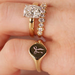 A close-up of a hand wearing three gold rings: a diamond engagement ring, a diamond-studded band, and a gold signet ring with a black oval face and a pink bow engraving.