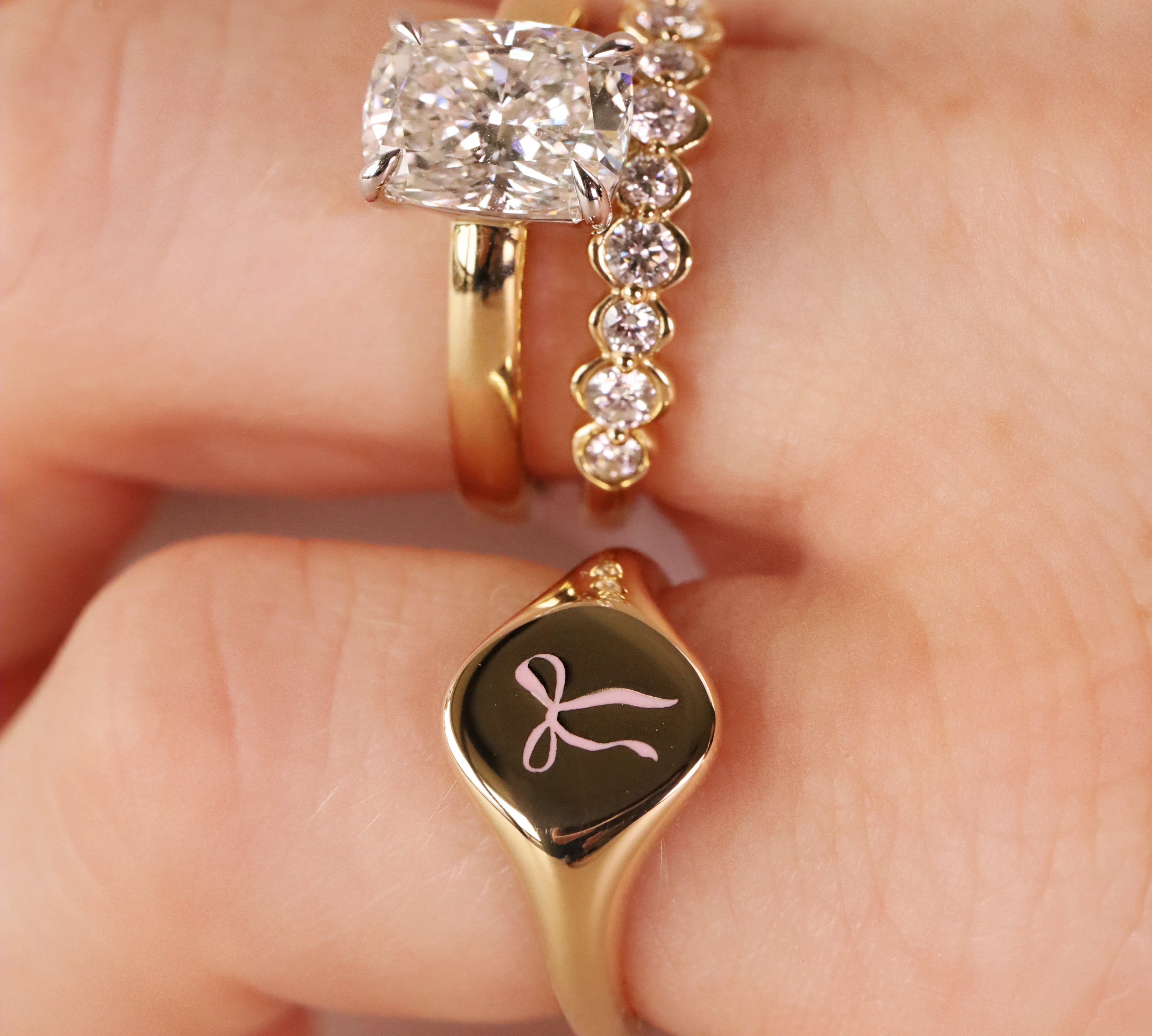 A close-up of a hand wearing three gold rings: a diamond engagement ring, a diamond-studded band, and a gold signet ring with a black oval face and a pink bow engraving.