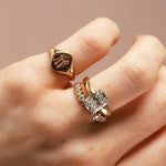 A close-up of a hand wearing three rings: a gold signet ring with a bow motif, a gold band with small clear stones, and a gold ring with a square clear gemstone and floral accents.