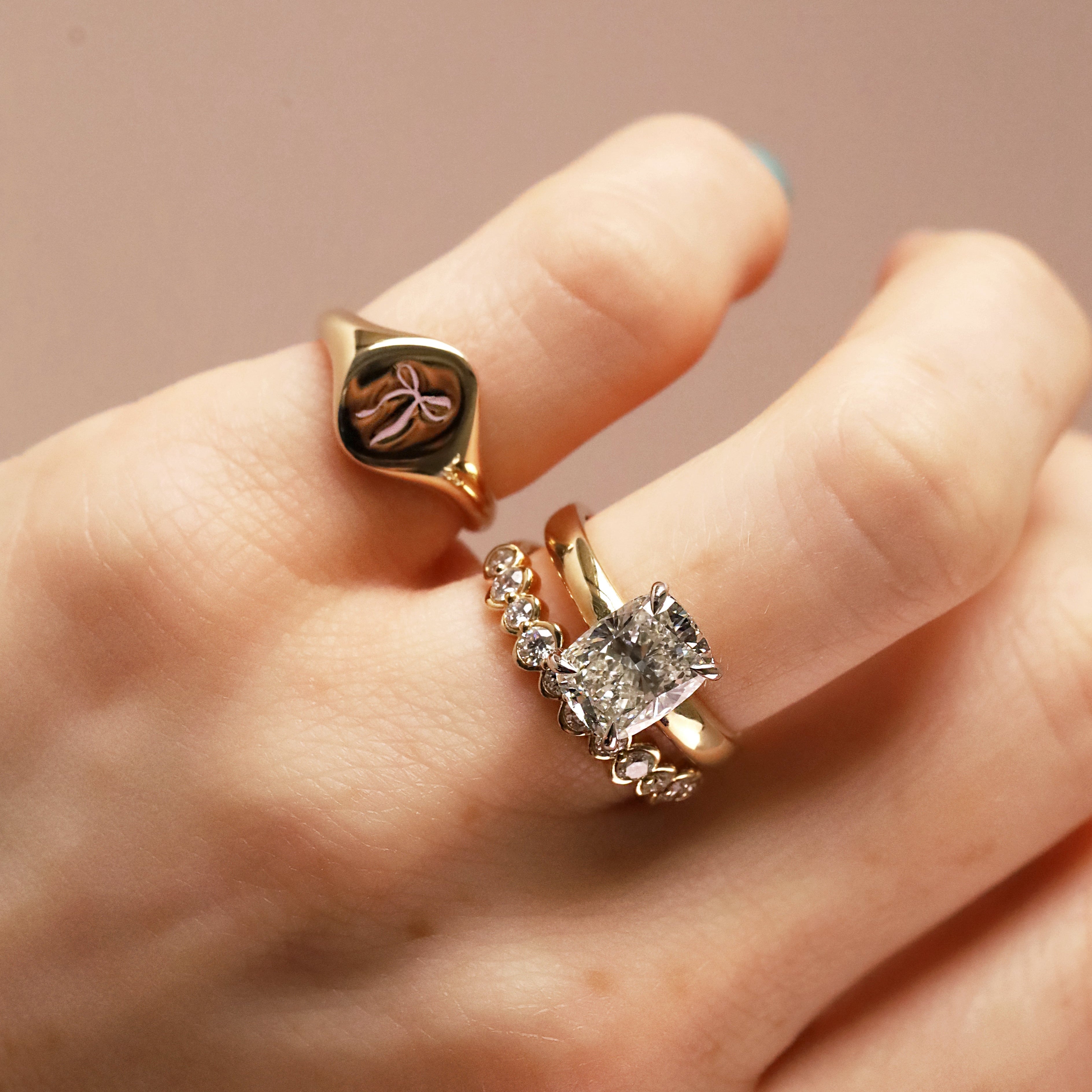 A close-up of a hand wearing three rings: a gold signet ring with a bow motif, a gold band with small clear stones, and a gold ring with a square clear gemstone and floral accents.