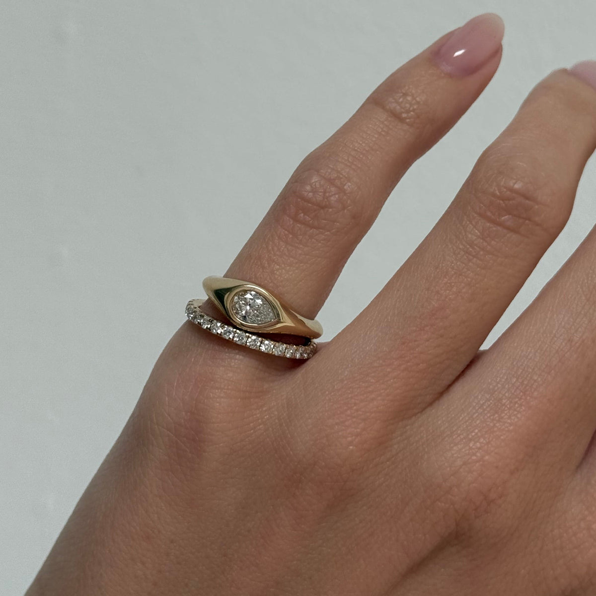 A close-up of a hand wearing two rings: a gold ring with a marquise-cut clear gemstone and a silver-toned pavé band ring, against a plain light gray background.