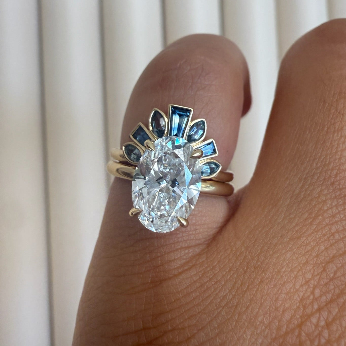 A close-up of a hand wearing a yellow gold ring with a large oval clear center stone and blue accent stones around the top, set against a background of white vertical blinds.