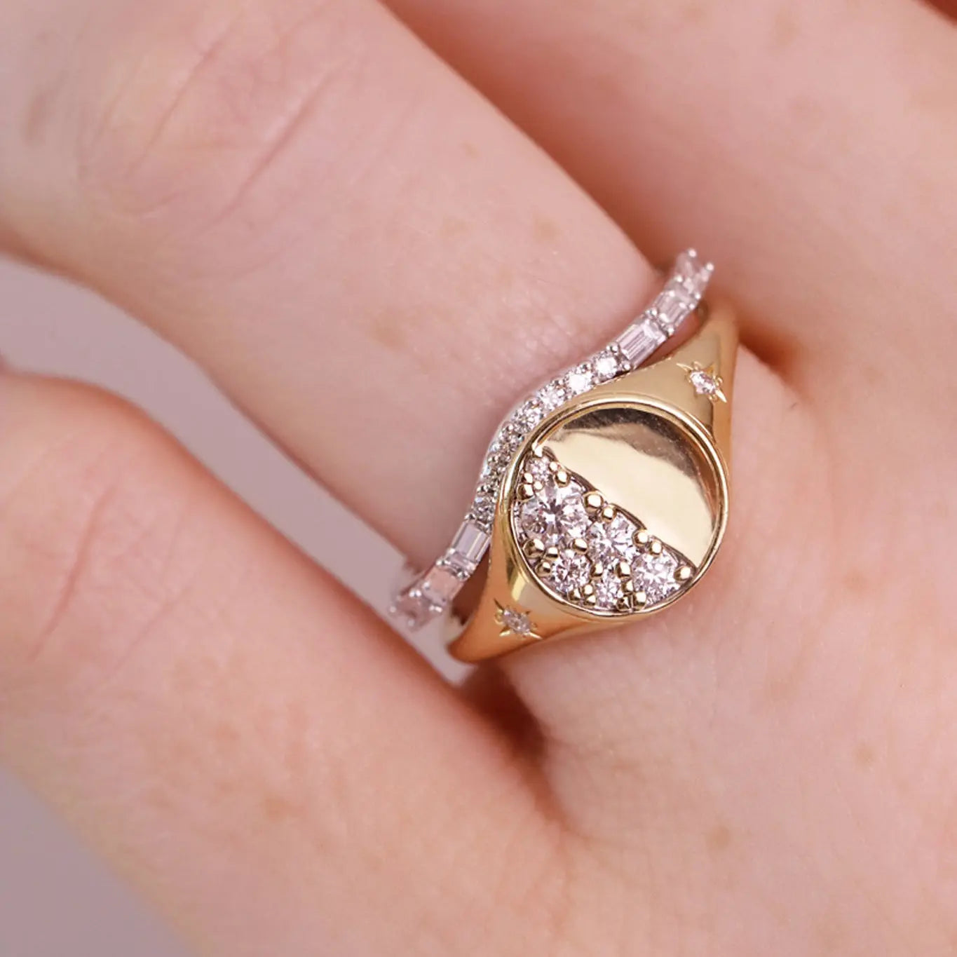 A close-up of a hand wearing a gold signet ring with a moon-phase design and pavé-set clear stones, alongside a thin silver band.