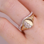 A close-up of a hand wearing a gold signet ring with a moon-phase design and pavé-set clear stones, alongside a thin silver band.