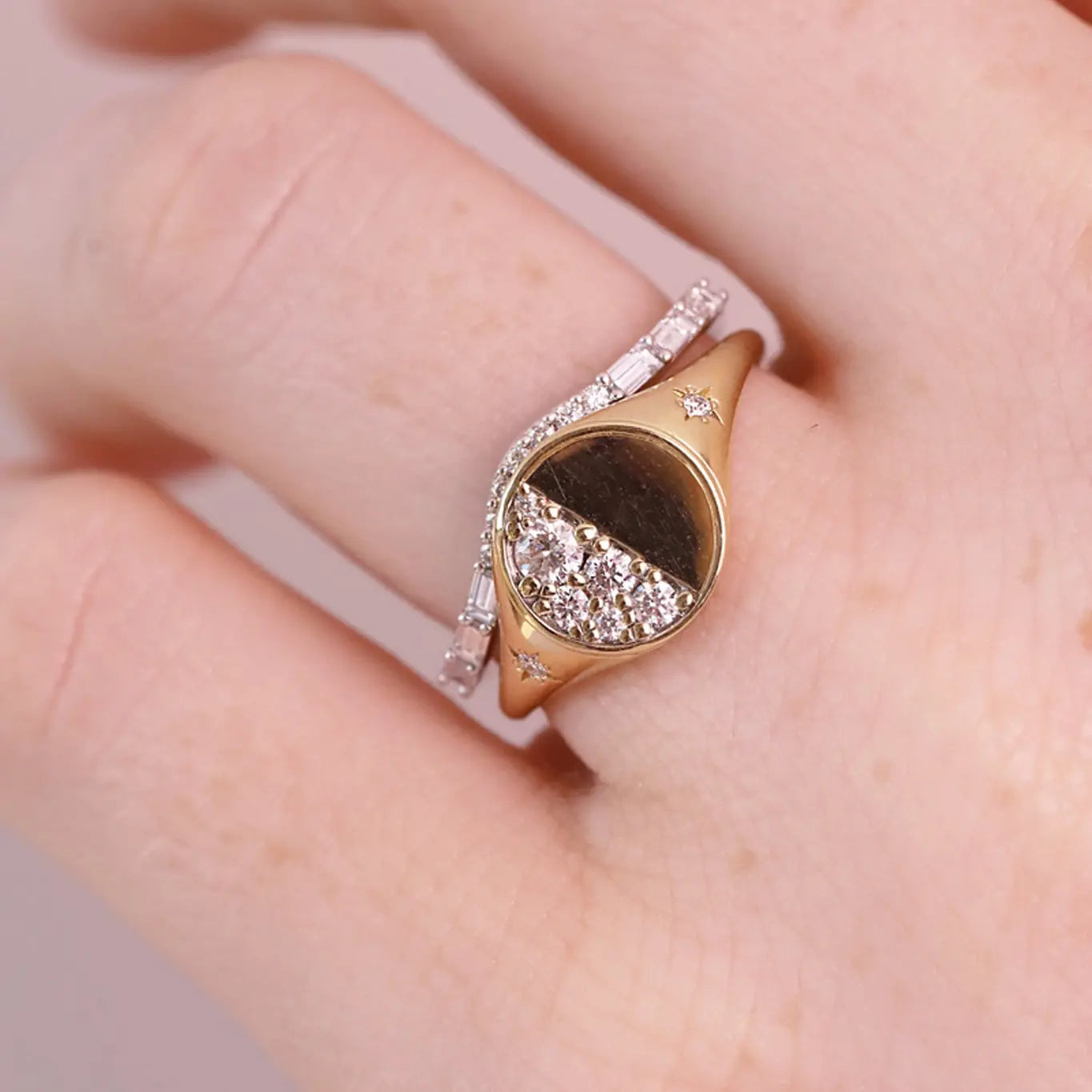 A close-up of a hand wearing a rose gold signet ring with a moon phase design set with clear stones, layered with a thin white diamond band.
