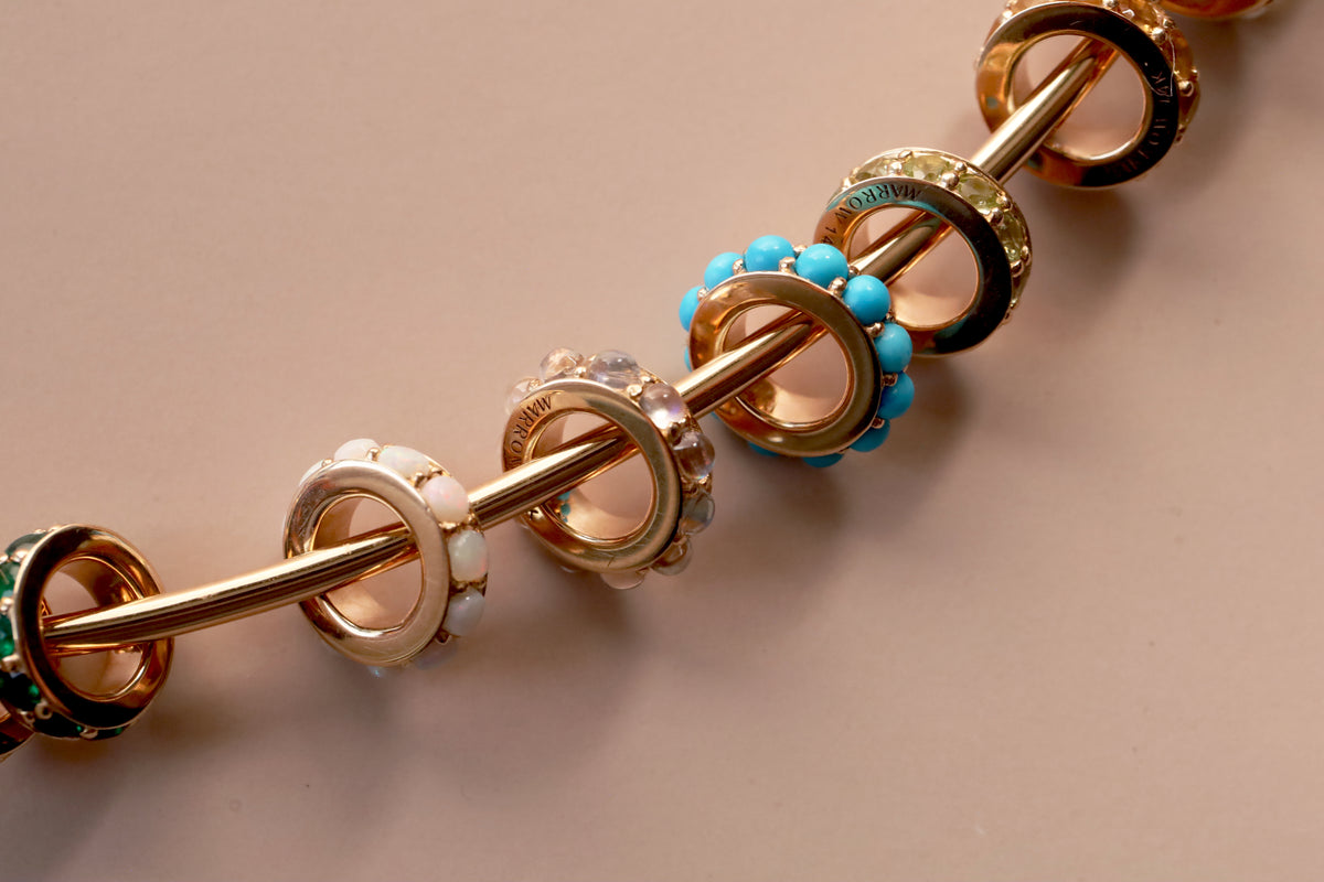 A close-up of a gold-toned necklace with multiple decorative charms in various gemstones, including turquoise, opals, emeralds, and moonstone, arranged on a solid beige background.
