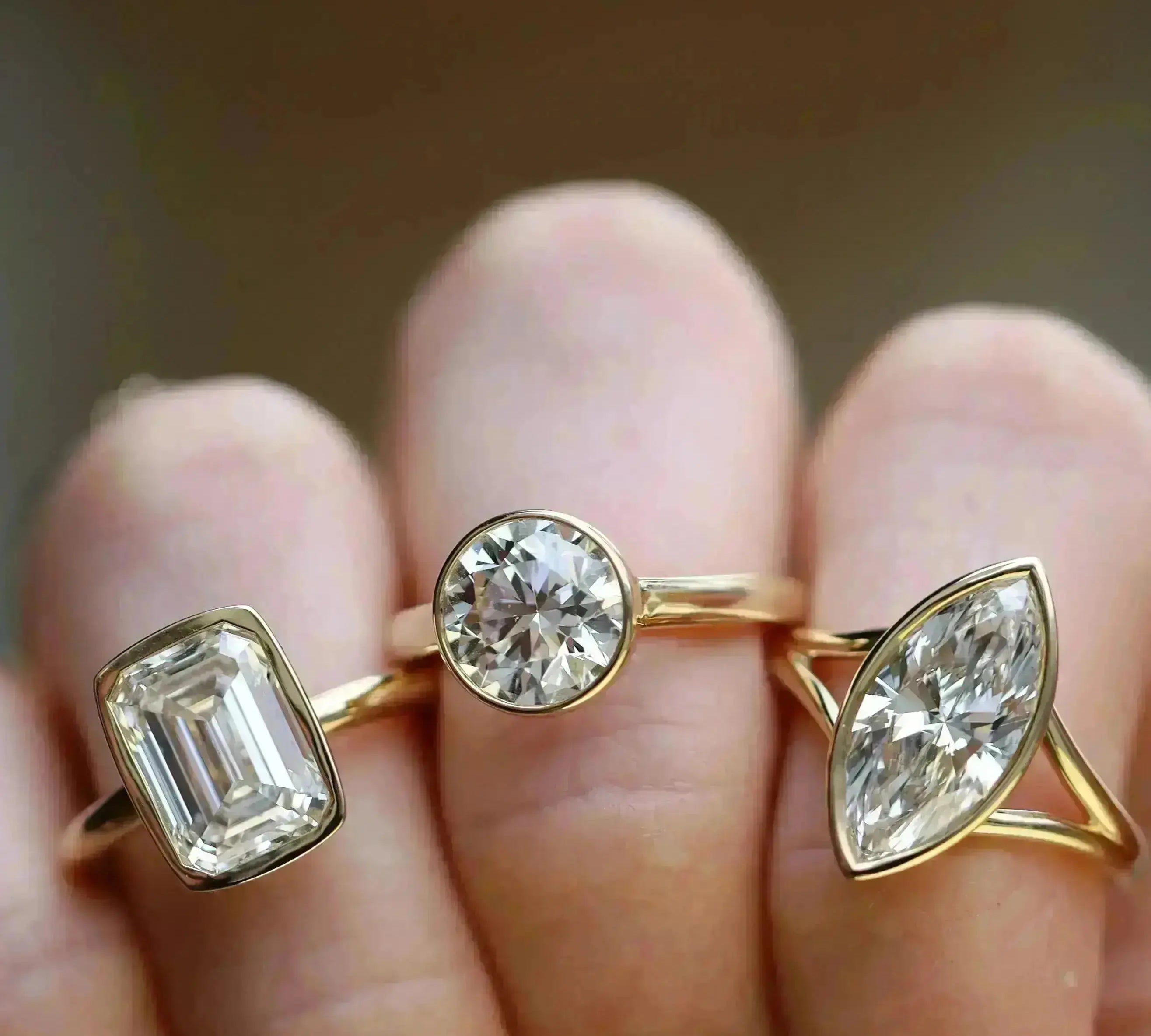 Close up of The Roxy Bezeled Emerald Cut Engagement Ring + The Tessa Bezel Round Solitaire Engagement Ring + The Colette Marquise Split Shank Engagement Ring