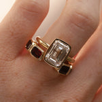 Open shank yellow gold and garnet band with subtle milgrain bezel detail stlyled on woman's hand with a emerald cut diamond engagement ring against neutral background