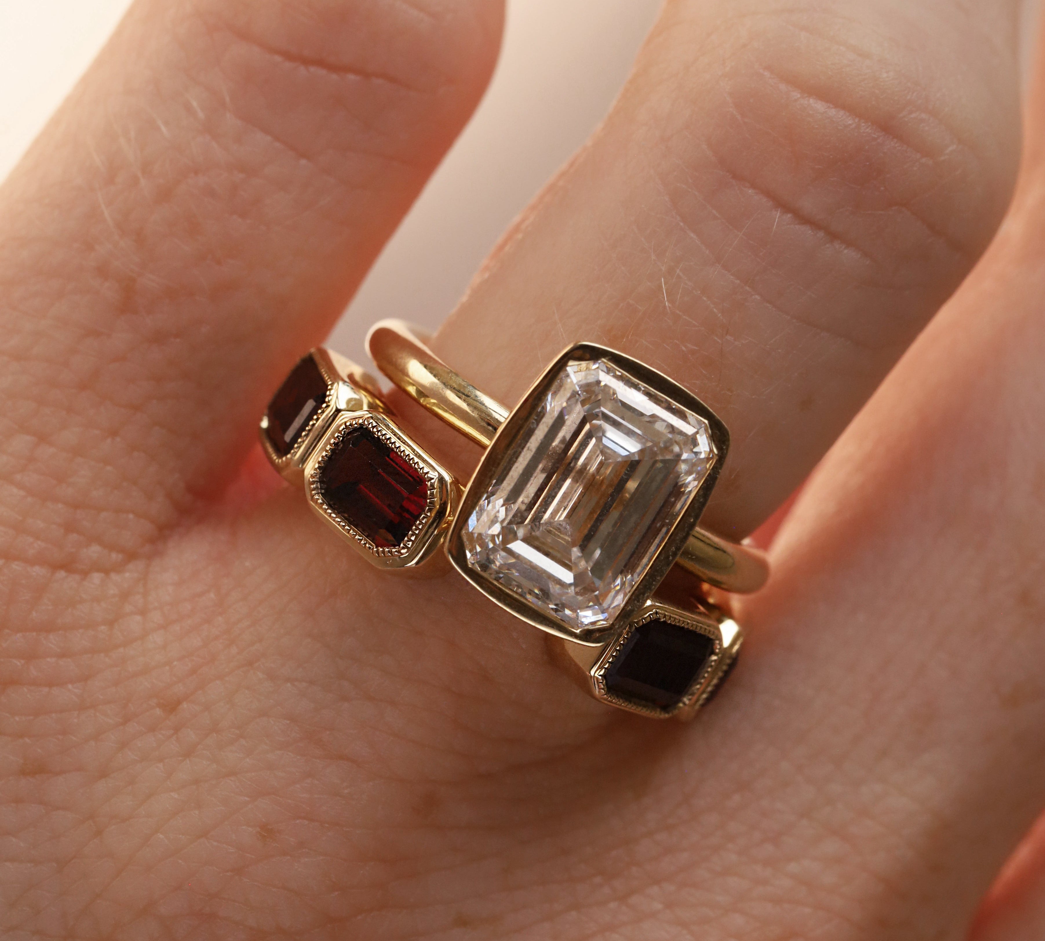 Open shank yellow gold and garnet band with subtle milgrain bezel detail stlyled on woman's hand with a emerald cut diamond engagement ring against neutral background