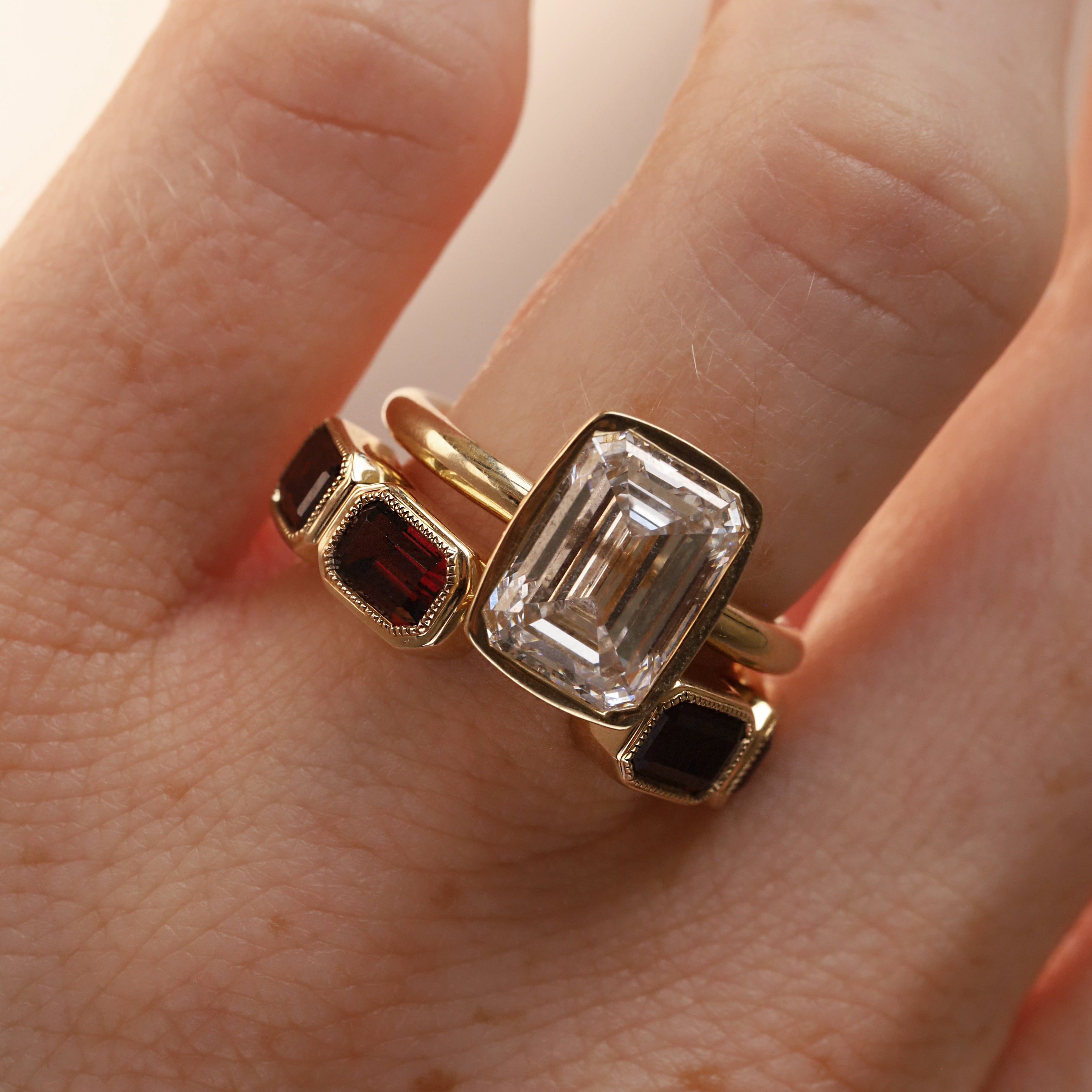 Open shank yellow gold and garnet band with subtle milgrain bezel detail stlyled on woman's hand with a emerald cut diamond engagement ring against neutral background