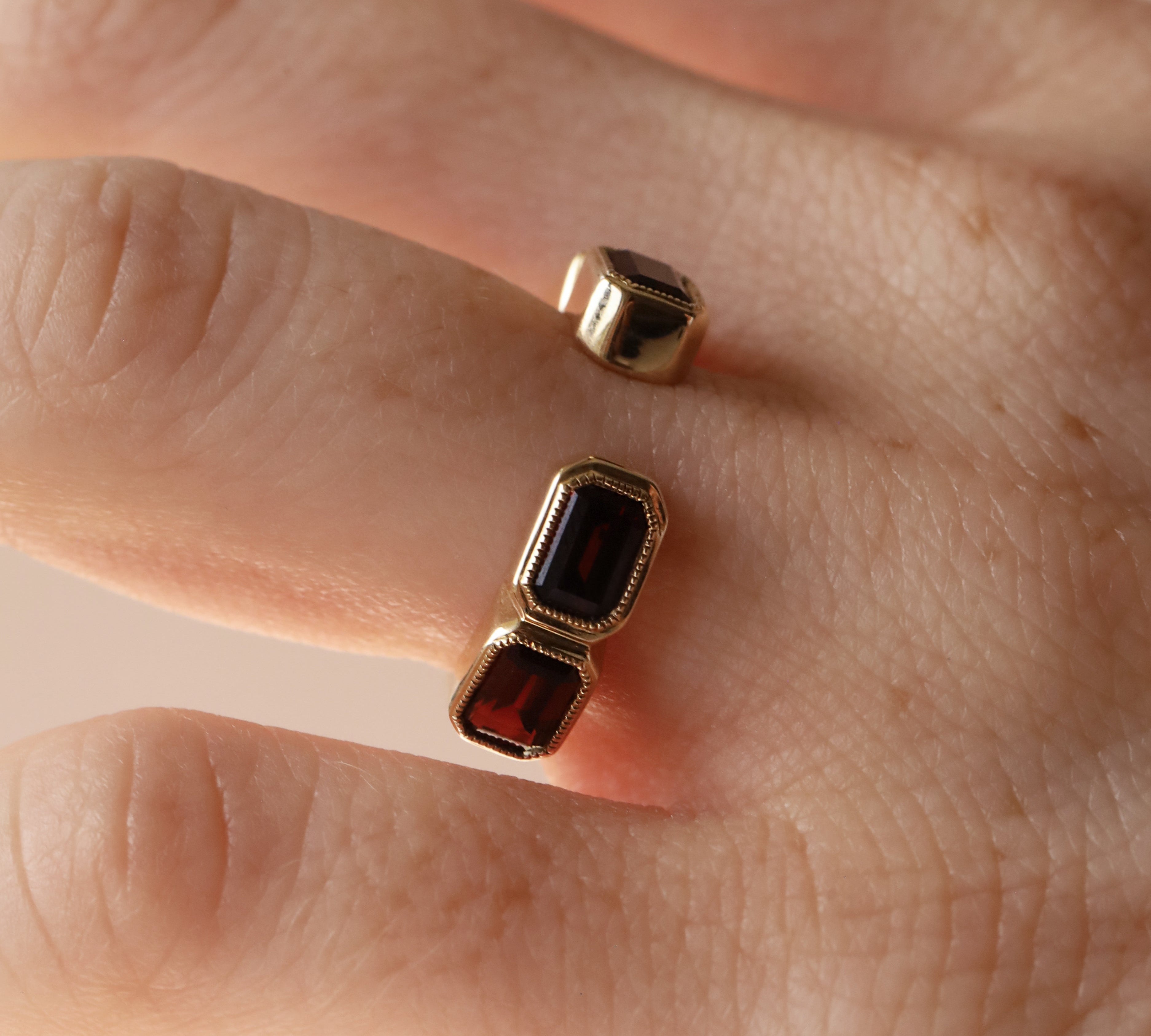Open shank yellow gold and garnet band with subtle milgrain bezel detail on woman's hand against neutral background