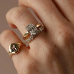 A close-up of a hand wearing three gold rings: a prominent engagement-style ring with a rectangular clear gemstone, a slim band with small clear stones, and a signet ring with a bow enamel design.