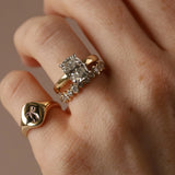 A close-up of a hand wearing three gold rings: a prominent engagement-style ring with a rectangular clear gemstone, a slim band with small clear stones, and a signet ring with a bow enamel design.