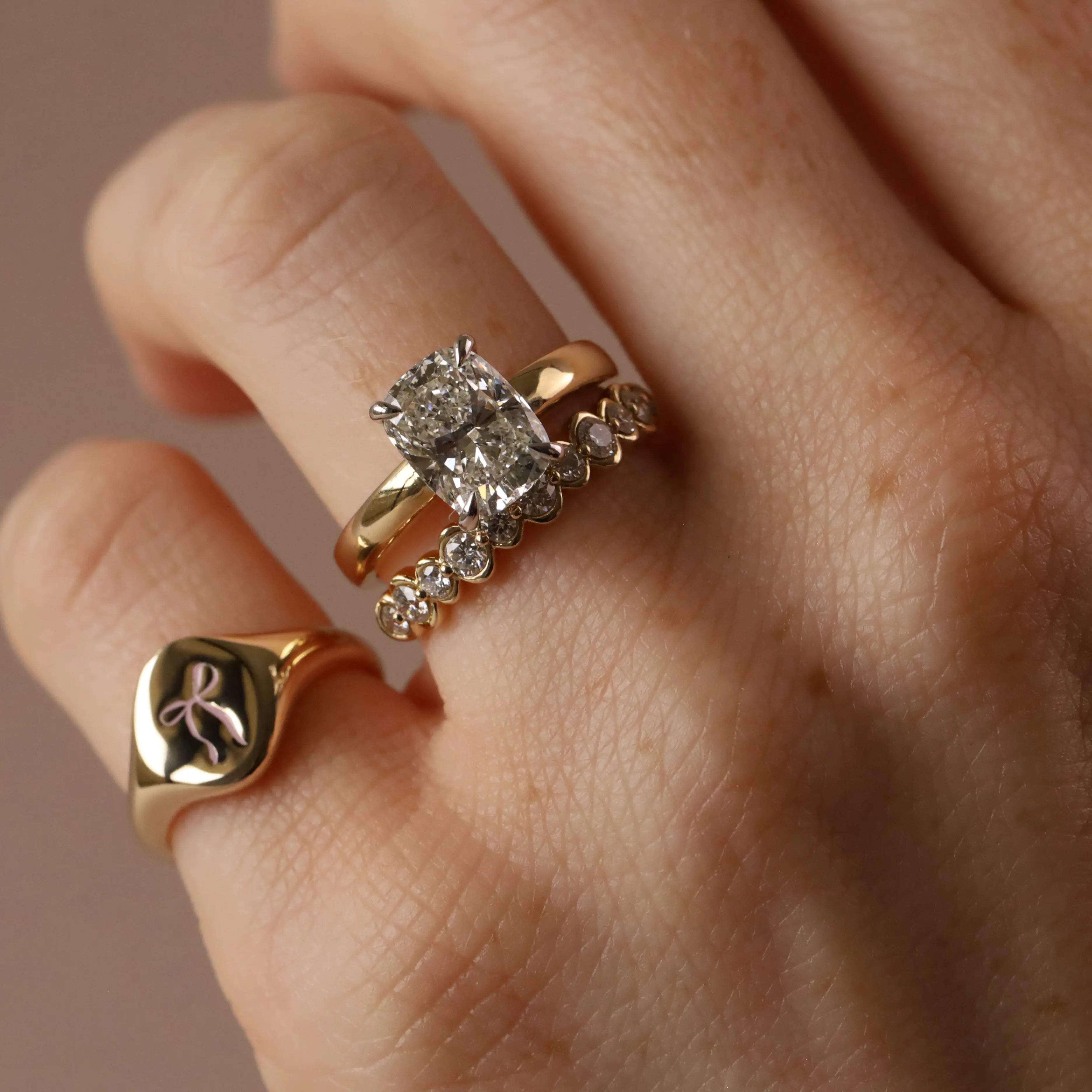 A close-up of a hand wearing three gold rings: a prominent engagement-style ring with a rectangular clear gemstone, a slim band with small clear stones, and a signet ring with a bow enamel design.