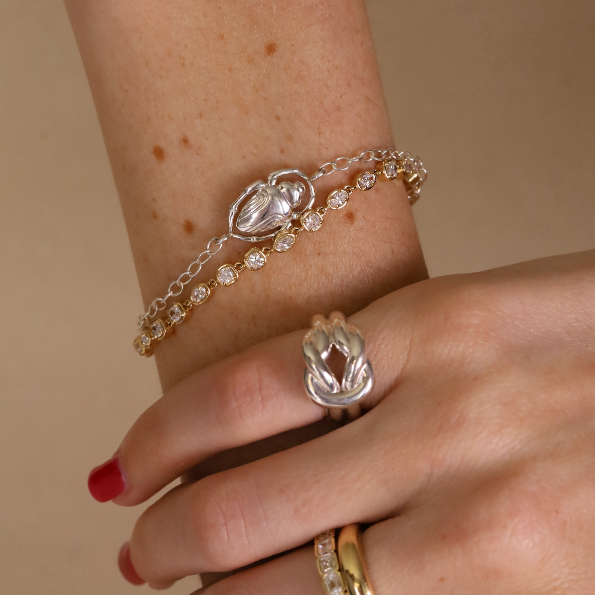 Close-up of a wrist wearing a silver bracelet with a clasp and two gold rings on a beige background.