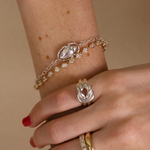Close-up of a wrist wearing a silver bracelet with a clasp and two gold rings on a beige background.
