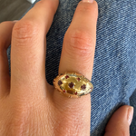 A gold oval ring with red gemstone accents worn on a finger, shown against blue denim fabric.