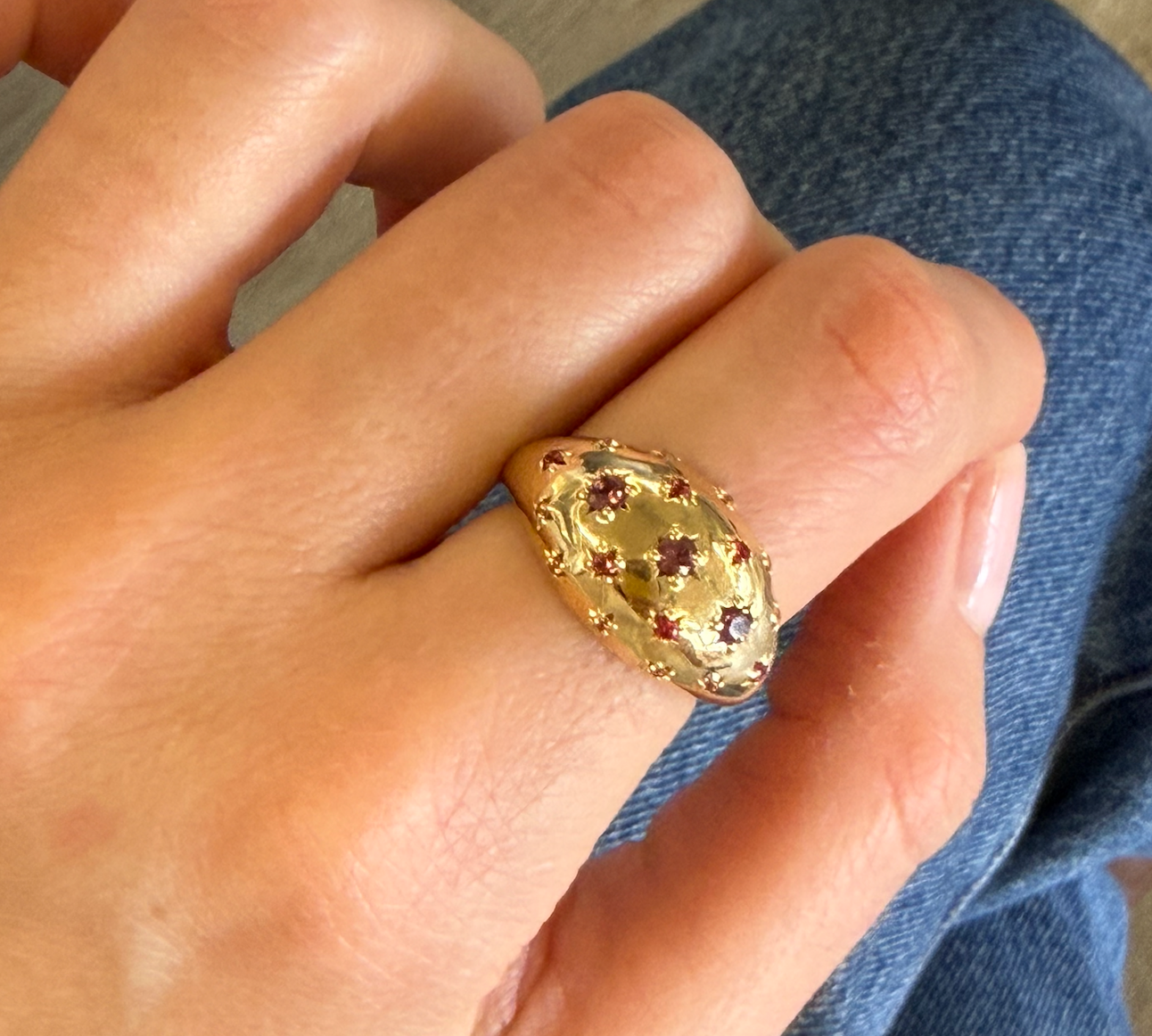 A gold ring with a textured, honeycomb-like oval face set with small red gemstones, worn on a person's finger against a background of blue denim jeans.