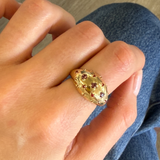 A gold ring with a textured, honeycomb-like oval face set with small red gemstones, worn on a person's finger against a background of blue denim jeans.