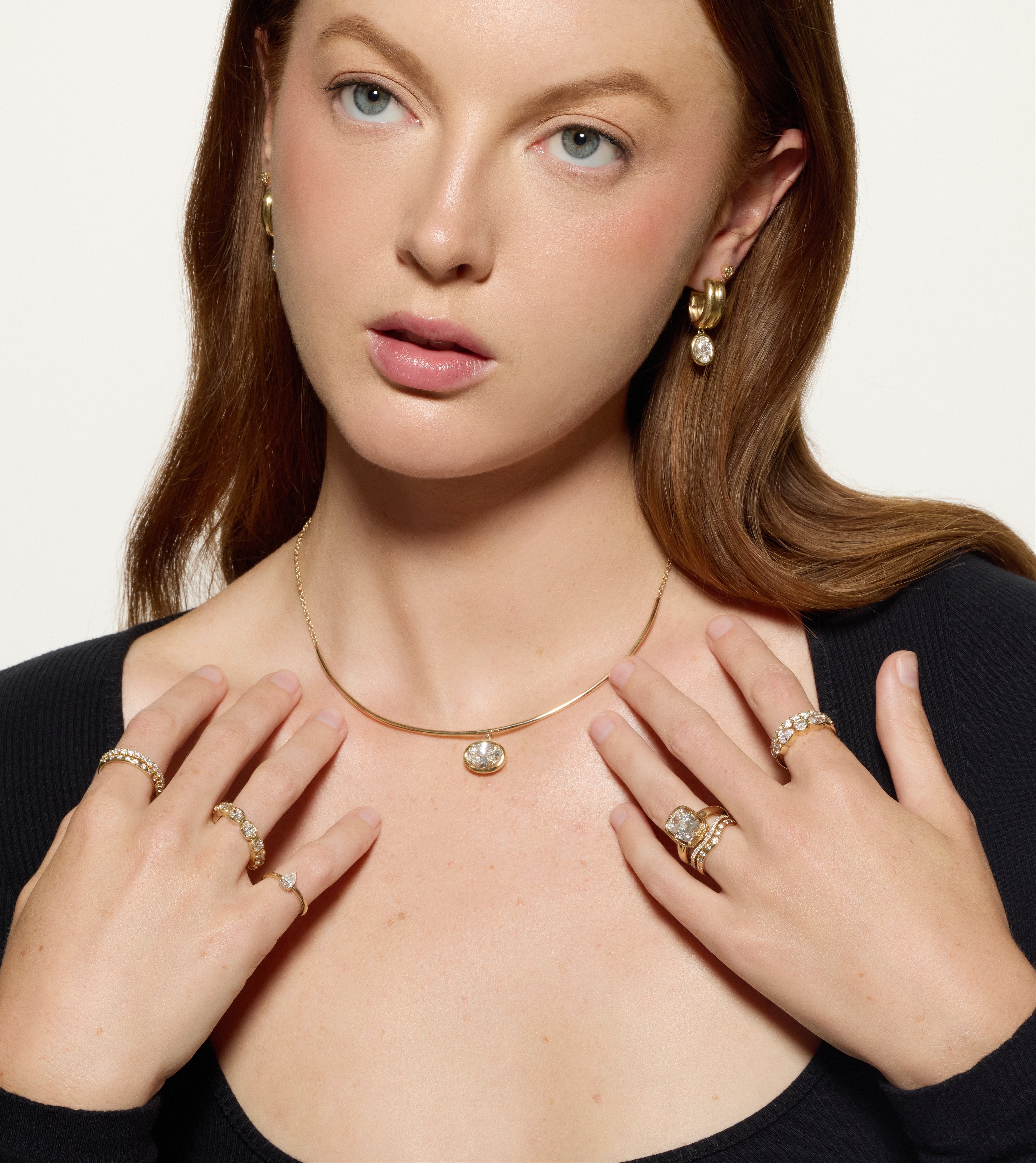 A close-up of a woman wearing a delicate gold necklace with a small oval pendant, along with matching gold rings and earrings, set against a clean off-white background.