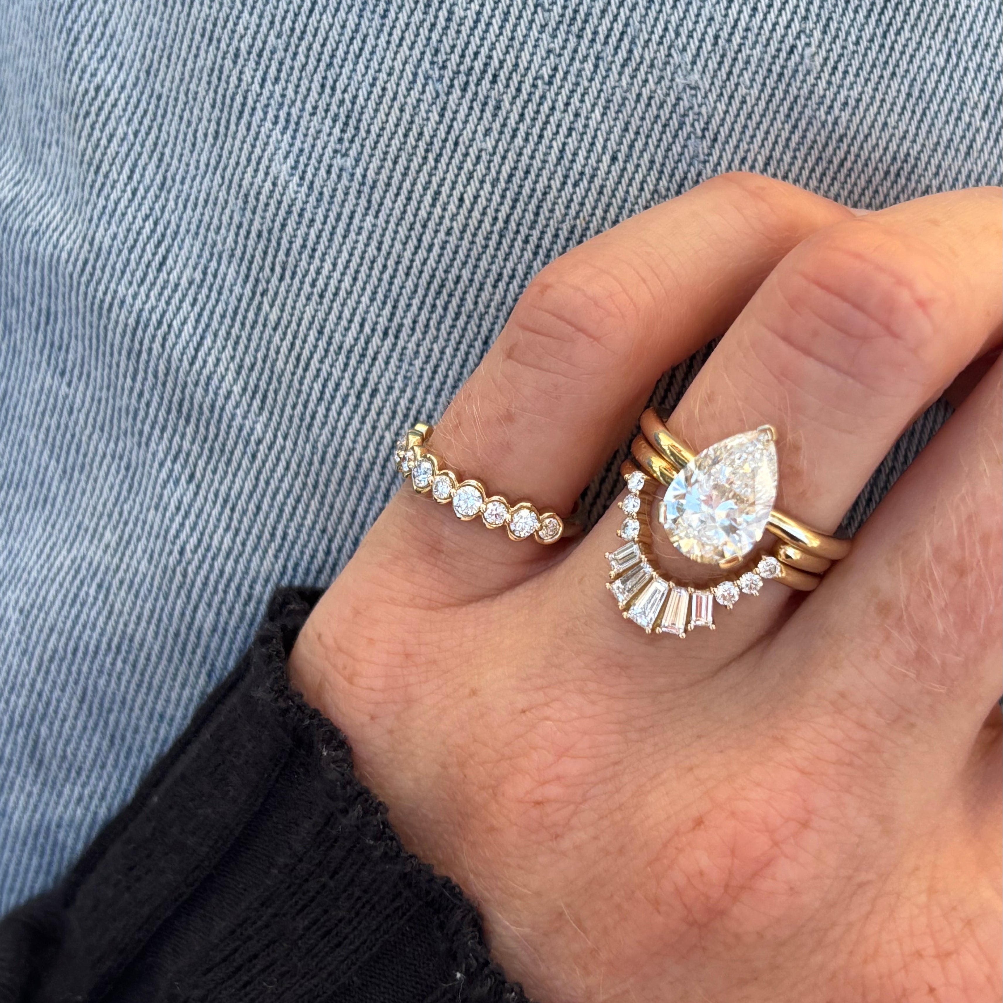 A close-up of a hand wearing a gold ring set featuring a pear-cut diamond center stone surrounded by baguette and round diamonds, alongside a thin diamond band on a light blue denim background.