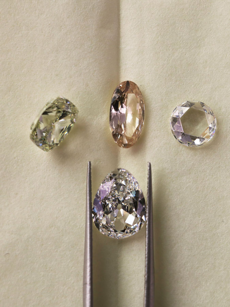 Four loose gemstones in different shapes and colors are arranged on a beige surface, with one oval pink stone and one round clear stone held in metal tweezers below.