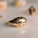 A gold teardrop-shaped ring with star-shaped engravings and small purple gemstones, photographed on a light gray surface with a soft-focus background.